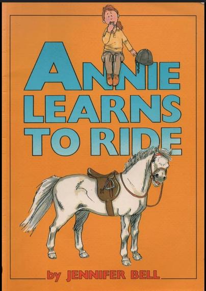 The front cover of Annie Learns To Ride by Jennifer Bell. The cover is plain orange with the title and author in light blue text. A nervous looking girl in jodhpurs holding her riding hat is sat on the Ns in Annie and beneath the title is a cheeky looking grey pony. 