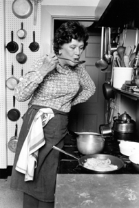 Julia Child standing at a stove sipping from a large cooking spoon.