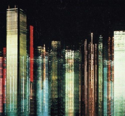 Stylized nighttime cityscape. Skyscrapers appear as vertical streaks of light against a dark background. The buildings are rendered in vibrant colors - yellows, reds, blues, and greens - creating an abstract, luminous representation of a city's energy and dynamism.