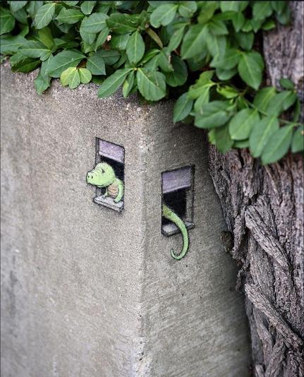 Streetart. A small green lizard (named Carina) was painted with crayon and a 3D effect on the corner of a low gray concrete wall of a plant bed. The mural is divided into two parts on the two walls of the corner. On the left side there is a small window with raised shutters, from which the lizard looks out. On the right-hand side, a little further down, there is another window from which the lizard's tail can be seen.
Title: 