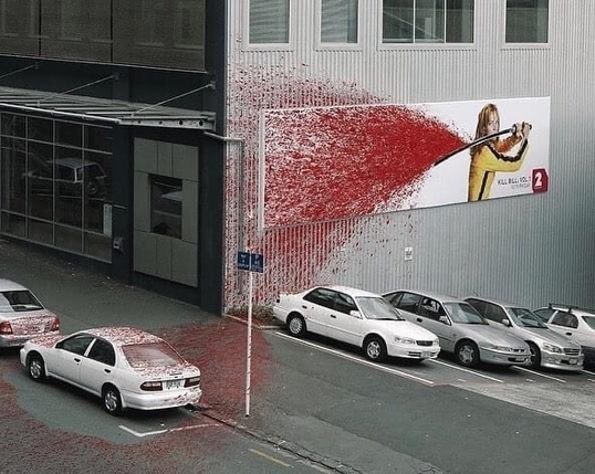 The focal point is a large billboard on the side of a building. It depicts a partial image of the film's protagonist in her iconic yellow jumpsuit, wielding a sword. The billboard is designed to make it appear as if the character has slashed the sword across the surface, creating a spray of red that extends beyond the billboard's boundaries.

This red spray appears to continue down the side of the building and onto the street below, covering part of a parked white car. The effect creates an illusion that the action on the billboard has impacted the real world.

The scene is set in what looks like a city parking area, with several white cars parked along the street. The contrast between the stark urban setting and the dramatic red spray makes the advertisement particularly eye-catching.

This advertisement exemplifies the bold, graphic style associated with the film, effectively capturing its themes of action and violence in a visually arresting way that integrates with its urban surroundings.

The image showcases an innovative approach to movie marketing, blurring the line between the advertisement and its environment to create a memorable and impactful display.