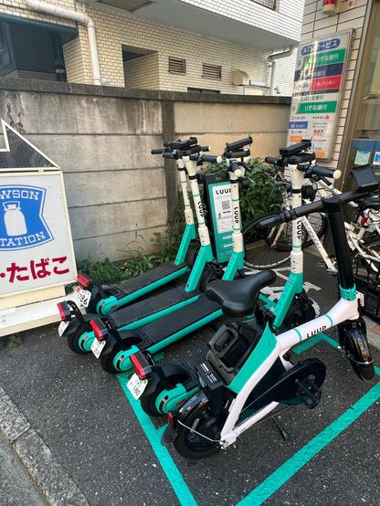 Four electric scooters and a small electric bike are parked outside a convenience store. The bikes and scooters are all coloured white and teal.