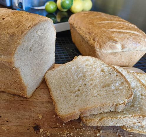 Two loaves of fresh white bread with a golden crust. The bread in the foreground is on a wooden cutting board and has three slices cut from the end. The cut pieces reveal that the loaf has a soft, well risen interior and a thin crunchy crust. 