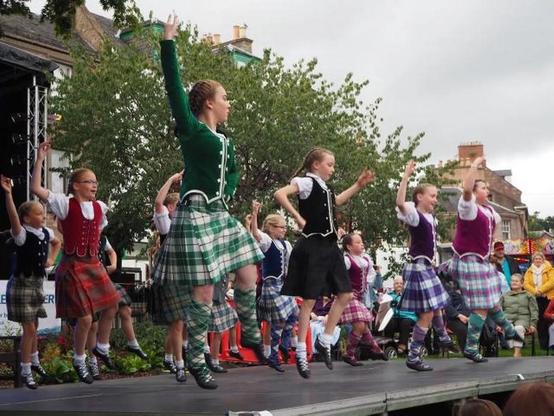 A group of young dancers performs on stage, wearing traditional Scottish attire, including kilts and vests. They are jumping and dancing energetically, with an audience watching in the background. Trees and buildings are visible in the scene, indicating an outdoor event