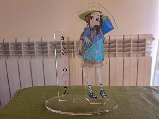 Foto de una figura de metacrilato de Takagi-san colocada en su soporte, sobre la mesa del comedor (con un calefactor detrás).

Takagi lleva ropa casual (una chaqueta azul, shorts beige y un sombrero amarillo), y extiende una mano hacia la cámara mientras lo otra la tiene sobre la nuca.

A su lado, en otro hueco del soporte, está colocada otra pieza de metacrilato con el título de la serie escrito en vertical, en japonés.