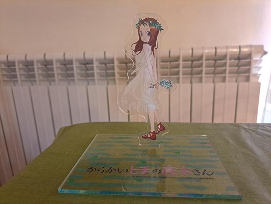 Otra figura de metacrilato de Takagi-san, esta vez llevando un vestido blanco de una pieza. Está dándonos la espalda, aunque con la cabeza vuelta hacia nosotros. Lleva una corona de hojas de olivo sobre la cabeza y unas ramas de olivo en las manos, que tiene juntas a su espalda.

El título de la serie está está vez en el mismo soporte, escrito horizontalmente.