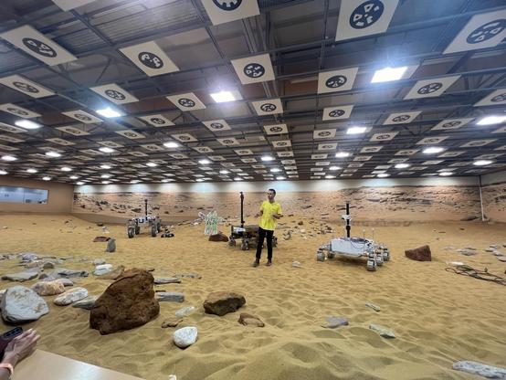 Presenter standing in a warehouse on sand with three Rovers.