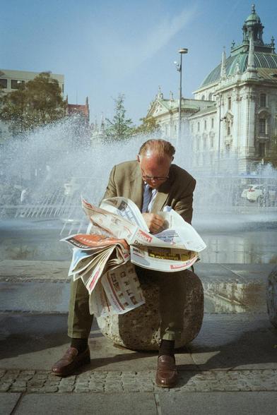 Photograph. A color photo of an elderly gentleman reading a newspaper. He is sitting on a large stone in a square with a fountain in the background. He is half bald, wears glasses and is dressed in a beige suit and brown leather shoes. The wind is blowing strongly from the side and he is struggling to tame the flying sheets of newspaper, but he is sitting engrossed in the daily paper. The wind can also be seen in the fountains spraying to the side in the background. The photographer was particularly interested in the flying newspaper. It seems impractical and outdated in this day and age, and the gentleman seems strange in his efforts to find peace and information while fighting against the wind.
Info: Ludwig Van Borkum is the pseudonym of Philipp Merz, a photographer from Munich who takes pictures of life on the city streets.