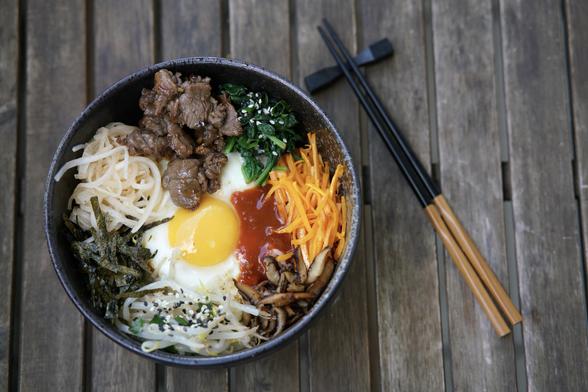 Un bol de bibimbap avec du bœuf, un œuf au plat, des carottes râpées, des algues, des épinards, des germes de soja et de la sauce, placé sur une table en bois à côté d'une paire de baguettes. 

A bowl of bibimbap with beef, fried egg, shredded carrots, seaweed, spinach, bean sprouts, and sauce, placed on a wooden table alongside a pair of chopsticks.