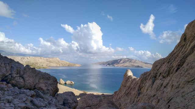 Photo of a small rocky valley ending in a beach in a small bay. The sky is blue with several large clouds.