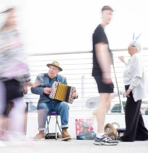 An older man sits playing an accordion while people pass by, blurred by a long camera exposure. A woman wearing blue rabbit ears watches on.