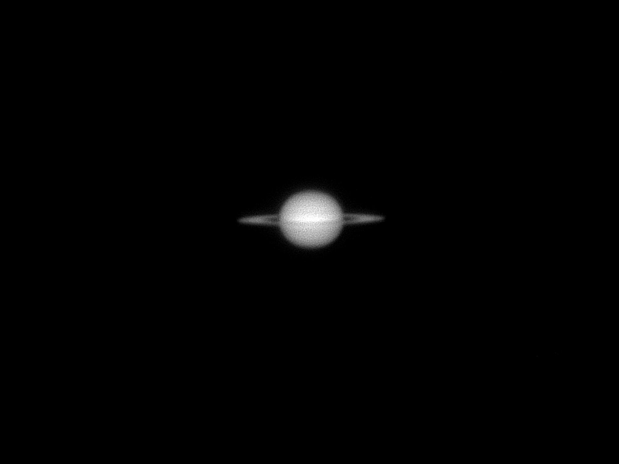 Telescopic view of Saturn - monochrome, north 'up'. Planet as a disc in the centre with some hints of horizontal bandings. Rings quite closed, there is a slither of black between the planet and the rings on either side. 