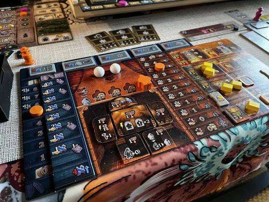 A tabletop board game is set up on a patterned tablecloth. The game board features various tokens in orange, yellow, and white, as well as tracks for scoring and resources. There are several cards and game pieces scattered around.