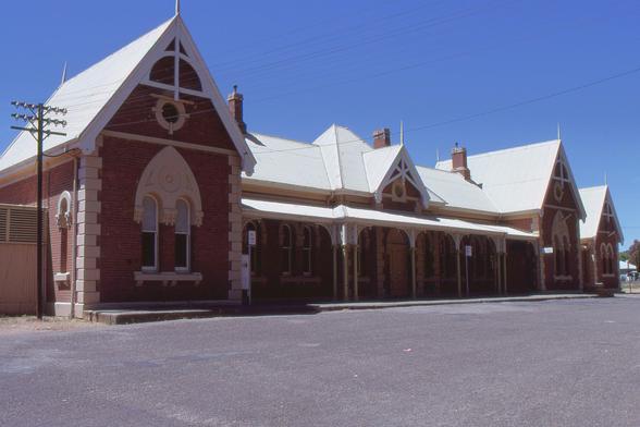 The front side of a brick and stone railway station building with corrugated iron peaked roof.  The building is of predominantly red brick construction with cream stone detailing on corners and detailing.  Windows are arched with ornate neo gothic styling.  At each end there is a transverse section with peaked roof and gable.  Connecting the two is a section that runs parallel to the platform, fronted with a verandah supported by metal posts with iron lacework.  A smaller central gable sits above a central entrance.  At the far end is another, perhaps detached room with another transverse peaked roof section.