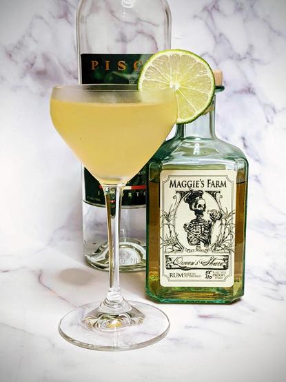 A bottle of pisco and a small bottle of Maggie's farm rum with a pale yellow cocktail with lime wheel in a Nick & Nora glass