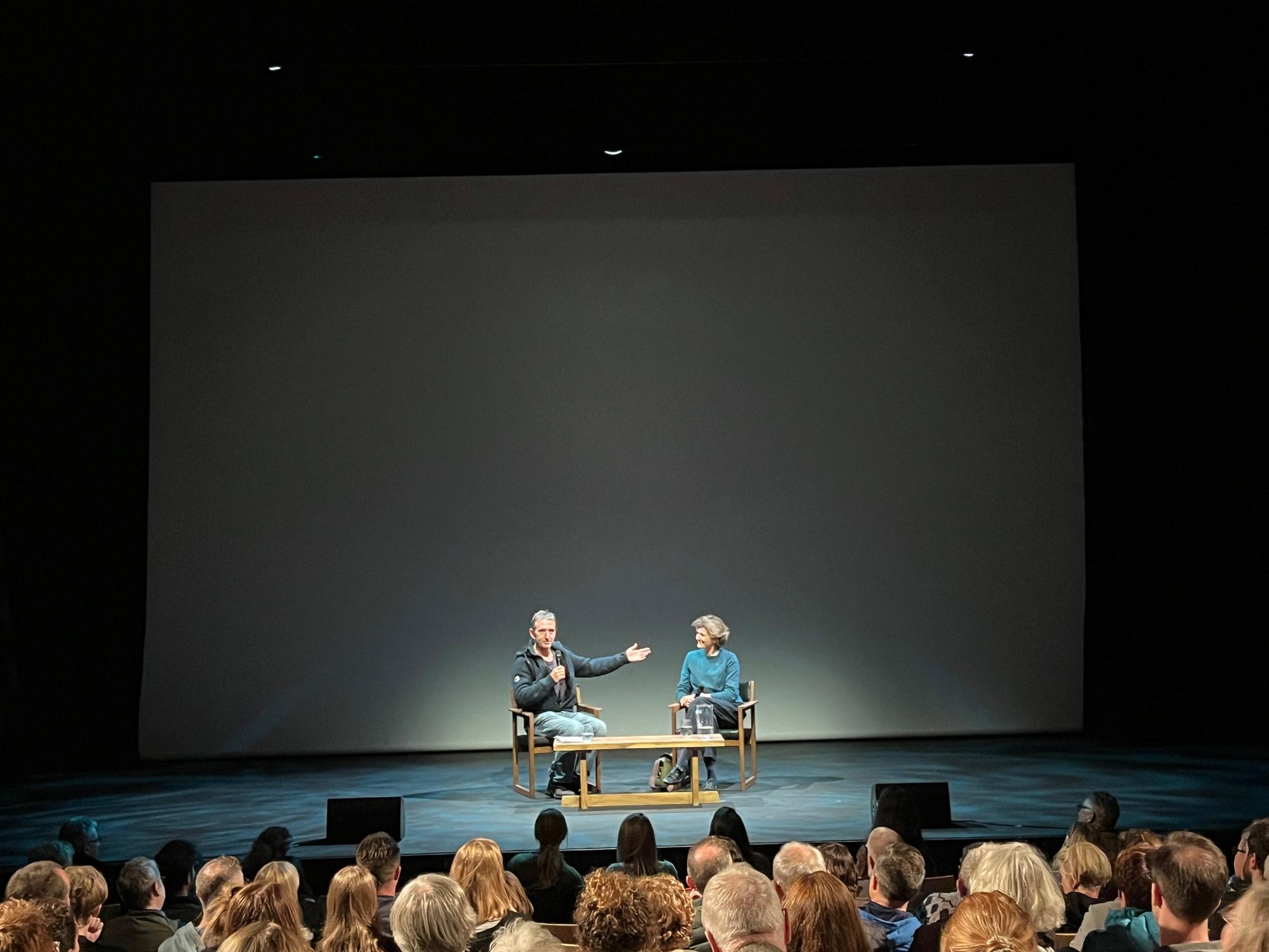 Man holding outstretched hand in direction of woman as he introduces her. The two people are sat on stools on an otherwise empty stage with a blank grey backdrop, as an audience looks on.