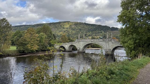 A stone bridge spans a river, surrounded by lush green trees and rolling hills under a partly cloudy sky. The scene is peaceful, with water reflecting the landscape.