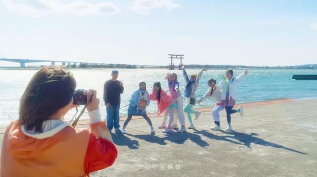 Imagen del mismo videoclip. Minami Natsu, componente de Candy Tune, de espaldas, fotografía con su cámara a sus seis compañeras (una de ellas con el perro en brazos) y el guía, frente al lago Hamana, con el toriii visible al fondo.