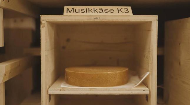 Das Foto zeigt eine Holzbox in der ein Laib Käse liegt. Auf der Box steht ein Schild mit der Aufschrift „Musikkäse K3“