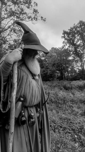 grey-scale photo of middle aged man with long grey beard and Gandalf style outfit with hat & staff standing in wooded marsh area looking down with shaded face