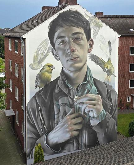 Streetartwall. An impressive and detailed portrait of a young man with a few greenfinches fluttering around his head was sprayed/painted on the exterior wall of a three-story apartment building. The color scheme is in shades of brown and yellow on a white background. The young brown-haired man is depicted up to his chest. He is wearing a patterned sweater and jacket and holds his hands in front of him as if he is waiting for a bird to perch on them. A yellow bird is already perched on his left shoulder, another is fluttering next to his right shoulder and more are flying in the background. The young man looks directly at the viewer. A mural designed in photorealistic quality that belongs to the master class in terms of color, light, detail and positive charisma.