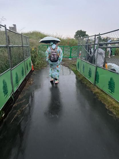 A woman wearing a pretty blue yukata and pink trainers carries and umbrella and backpack as she walks down a path. 