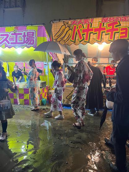 Three woman splash through puddles in front of food booths wearing yukata. One is wearing trainers while the other two are in the slim geta sandals without socks. 