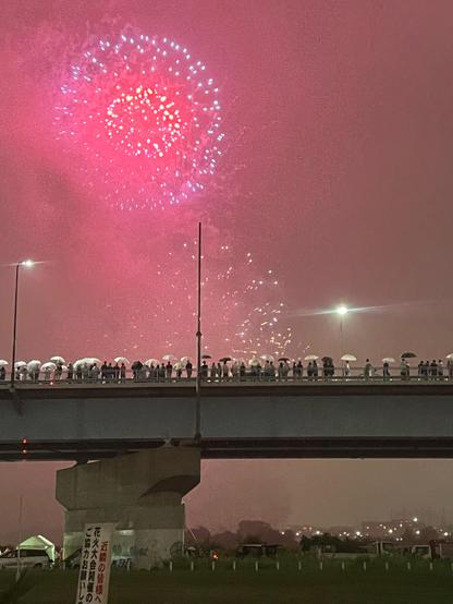 Red and purple fireworks explode in a circle about a bridge lined with people standing with umbrellas. 