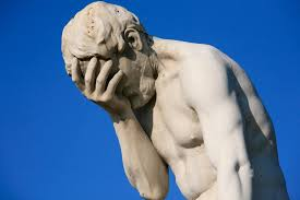 Image of a 'classical' style statue of a male human figure against a blue sky background. 
The figure is facepalming