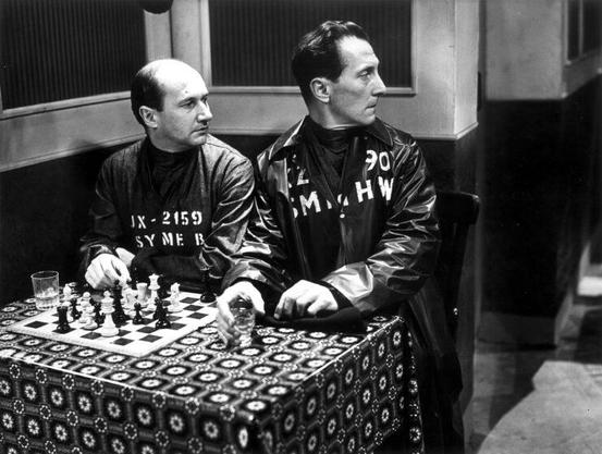 In a scene from the 1954 BBC production of 1984, Donald Pleasence and Peter Cushing sit side by side behind a chess set at a restaurant table.