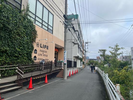 A building partially covered by ivy is on the left, with a fairly subtle sign on the outside wood wall saying “Life” and 2F for fashion and lifestyle, 1F for supermarket. The path next to it can be used by cars (maybe for access) but is clearly more suited for people and bikes. There are no cars in sight. 