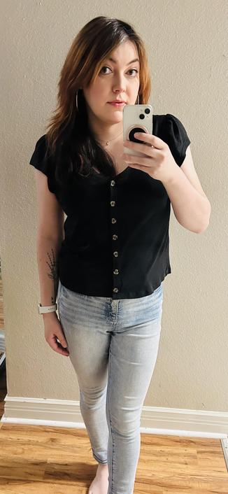 A woman (me) taking a photo in a mirror that is larger than the frame. I’m wearing a black short sleeve button up top with a slight puff sleeve and light wash denim pants. I’m also wearing large gold hoop earrings and small teardrop-shaped gold pendant necklace on a thin chain