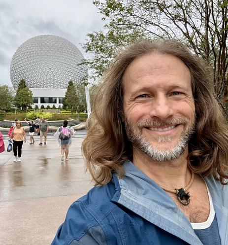 A man in a blue rain coat stands in front of Spaceship Earth. A handful of other guests walk by in the background. 