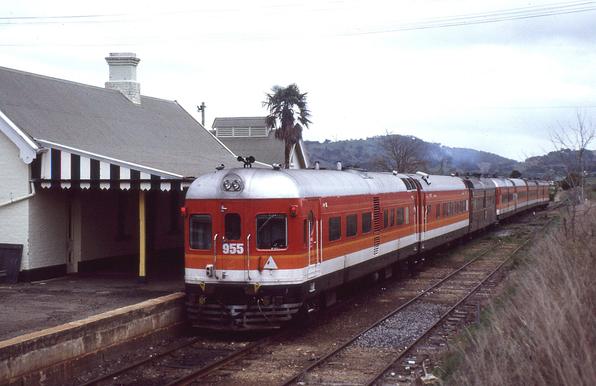 A DEB set train departs a rural passenger station.  The train is a 7-car streamlined diesel multiple unit with most cars painted in Candy livery but the third car from this end is in Indian Red livery.  The single platform station is at left.  There is just one track to the right of the train.