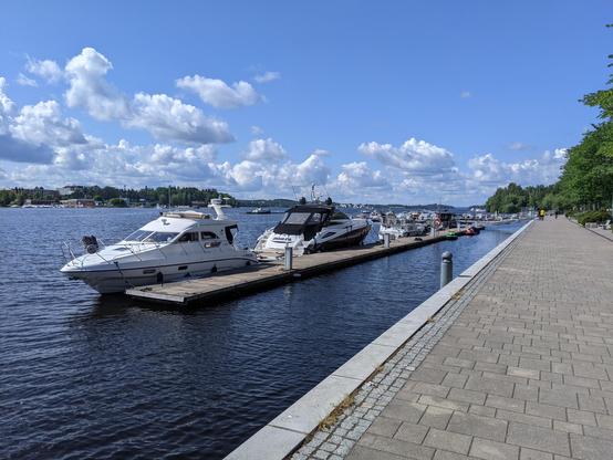 Des petits bateaux de plaisance amarrés à un ponton sur le lac de Tampere. À droite un quai piéton pavé bordé d'arbres, à gauche le lac, on aperçoit la rive d'en face au loin. Le ciel est bleu avec des nuages blancs.