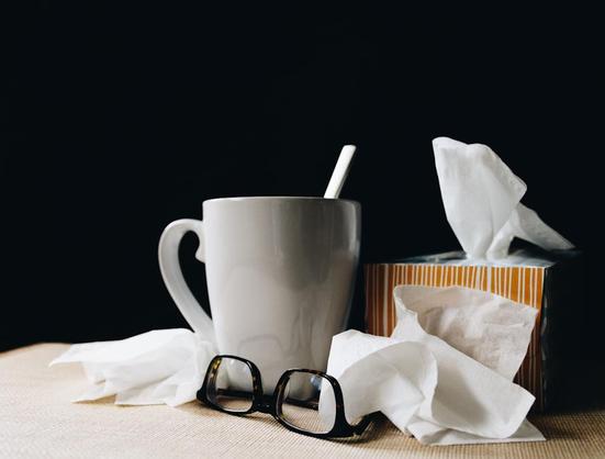 White mug with a spoon, a striped tissue box, crumpled tissues, and glasses on a table against a dark background.