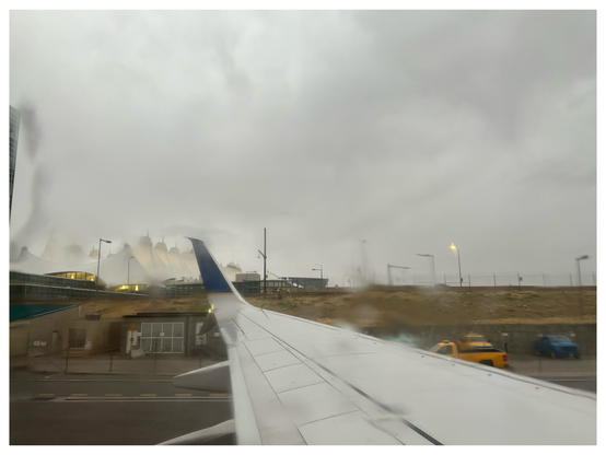 Photo with a white border around it showing a view of the Denver International Airport from a window seat showing the airplane wing, the runway, service vehicles, and the airport’s distinctive white tents. The atmosphere is misty and a golden hour light breaks through the clouds to give the scene a warm light.