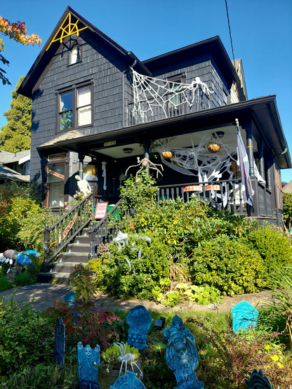 On the first Monday of October, with clear blue skies beyond, a Victorian house painted black is festooned with Halloween decorations of spiders, skeletons, spider webs, headstones, and ghosts.