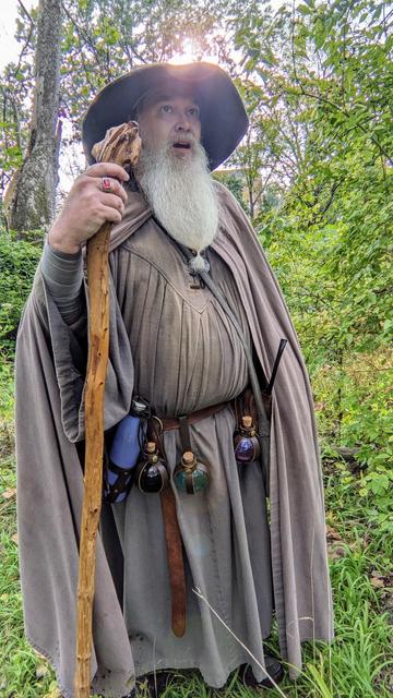 middle aged man with long grey beard standing in green wooded clearing wearing Gandalf style outfit with hatr & staff with amazed expression as sun rises over his hat