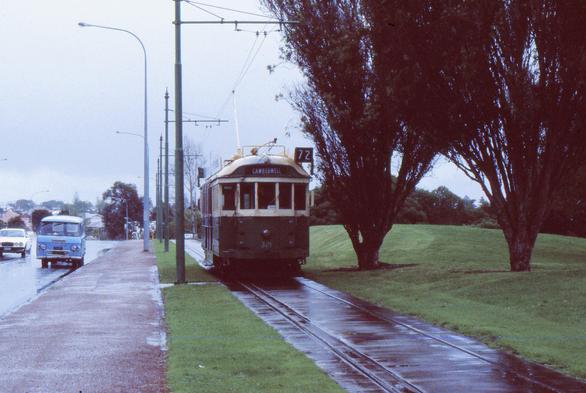 On a very wet, heavily overcast day, a Melbourne W2 class tram running on a single track dual gauge tramway set in concrete in parkland and running a along the side of a road.  The tram is displaying route 72 with a destination of Camberwell.