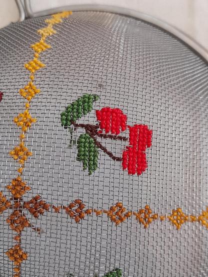 Cross stitch on a metal sieve: a yellow geometric pattern and cherries.