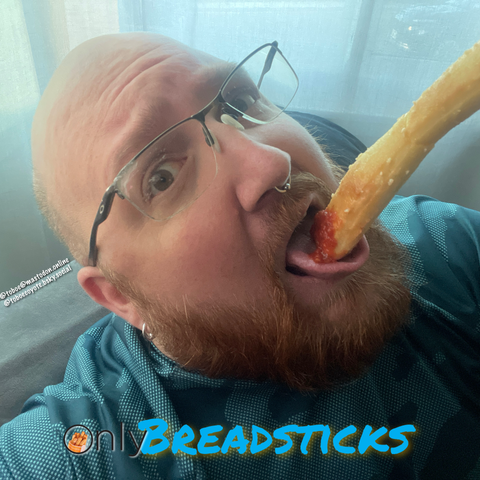 A selfie of me sitting in a chair against curtains. I'm holding a breadsticks dipped in marinara sauce suggestively onto my tongue, and looking seductively into the camera. A parody of the OnlyFans logo is at the bottom, reading OnlyBreadsticks, and an image of breadsticks appears in the letter O where the lock icon would be.