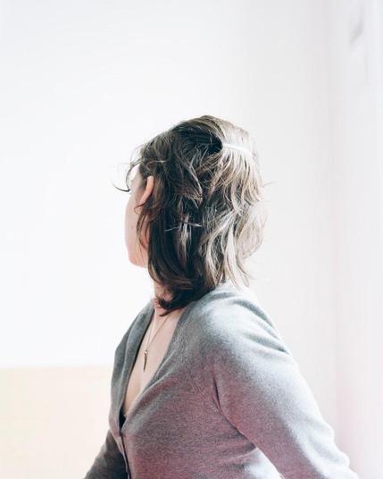 a woman with brown hair & grey, long sleeved sweater in a white room — her position mirrors the one of the woman in gerhard richter’s photorealistic painting “betty” (see above) — subject of photo & photographer unknown