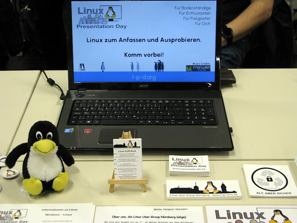 Bild: Ein Laptop mit dem Reklamevideo der LUG Reutlingen zum Linux Präsentationstag. Links davor ein kleiner Tux und mit unseren Visitenkarten.