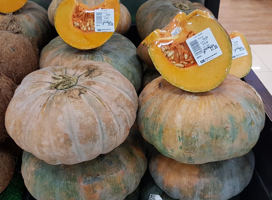 Photo of pumpkin-like squash stacked up at a market stall. They're about volleyball sized, a little flattened, a mix of dark green and chestnut/orange, with a dusty bloom. A few squash are cut up & wrapped in plastic so people can buy 1/4 of a squash.