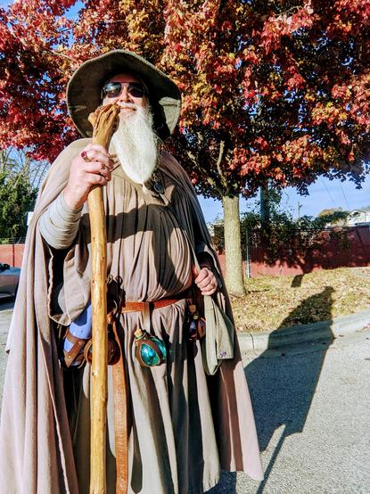 Middle aged man with long grey beard and sunglasses wearing a Gandalf style outfit stands smiling into the sun in front of red fall foliage