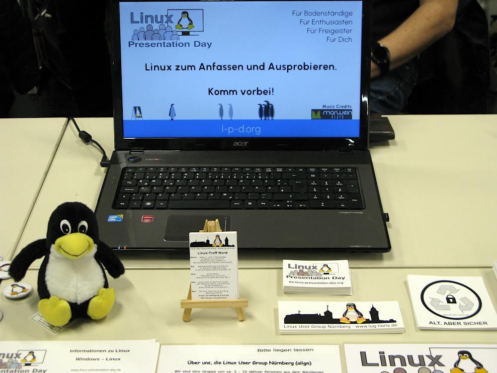 Foto: In der Bildmitte ein altes Notebook, vorne links der kleine Linux-Pinguin, in der Mitte unsere Visitenkarten und rechts davon noch einige Aufkleber. Das Ganze befindet sich auf einem Tisch.