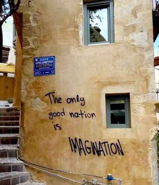 Graffiti on a weathered wall in Chania, Crete, Greece, reading "The only good nation is IMAGINATION&quot; in bold black letters. The phrase is creatively written across the textured surface of the ochre-colored building, situated near a staircase <br />leading upward. A blue street sign in Greek, labeled &quot;Οδός Δούλος Δημητρίου&quot; (Doukos Dimitriou Street), is mounted above the graffiti. The scene blends urban art with the historic charm of the neighborhood.