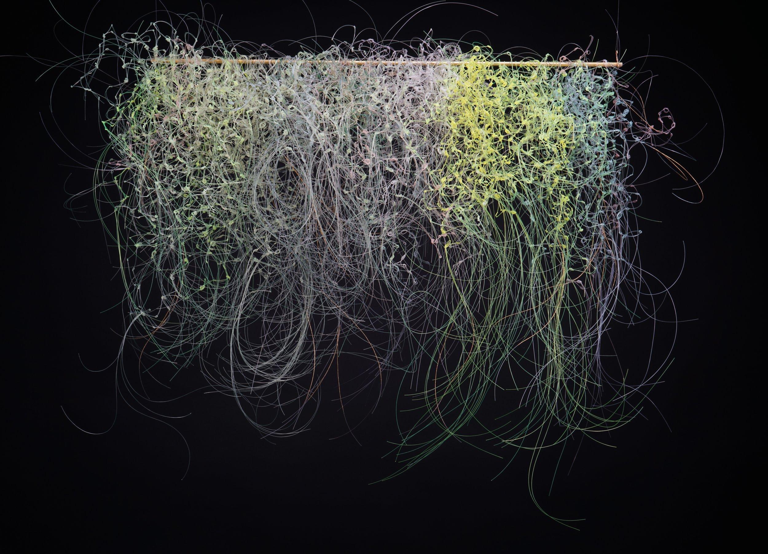 Hanging horizontal wall sculpture of thin green, white, and purple fibers cascading downward in clusters, with the ends swirling upwards