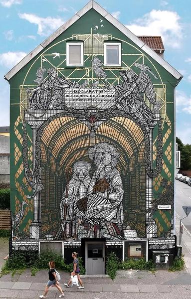 Streetartwall. A large (32 m x 10 m) and impressive mural with ornamentally drawn animals and the theme of human rights was installed on the outside wall of a two-storey building. The background is dark green, the mural itself is black and white. As with a mosaic, many individual parts were designed to create a detailed mural that looks like an old church window. The French artists were inspired by the "Universal Declaration of Human Rights" of 1789
and its text, which shows an engraving with two angels on a triumphal arch. It shows the frame of the original engraving, but reinterpreted: The angels hover over arches and porticoes on either side, symbolizing the moral pillars. The text reads as follows: "Déclaration des Droits de l'Homme et du Citoyen". At the center of the composition are two figures: a monkey and a bird, standing against the backdrop of the majestic library of Trinity College Dublin. They symbolize guardians and embody wisdom and knowledge. Power and intellect only take on their true meaning when they are guided by reason and ethics.
Info: The theme was the 17 UN Sustainable Development Goals. The mural illustrates goal 16 in its own way: Peace, justice and strong institutions.